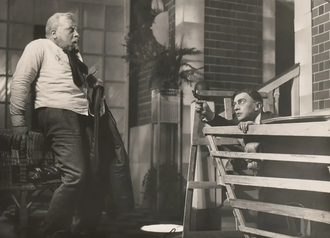 Szyfman reżyser teatralny uchwycony w dramatycznej scenie. Mężczyzna z pistoletem celuje zza drewnianej konstrukcji, podczas gdy drugi, starszy mężczyzna, reaguje z zaskoczeniem.