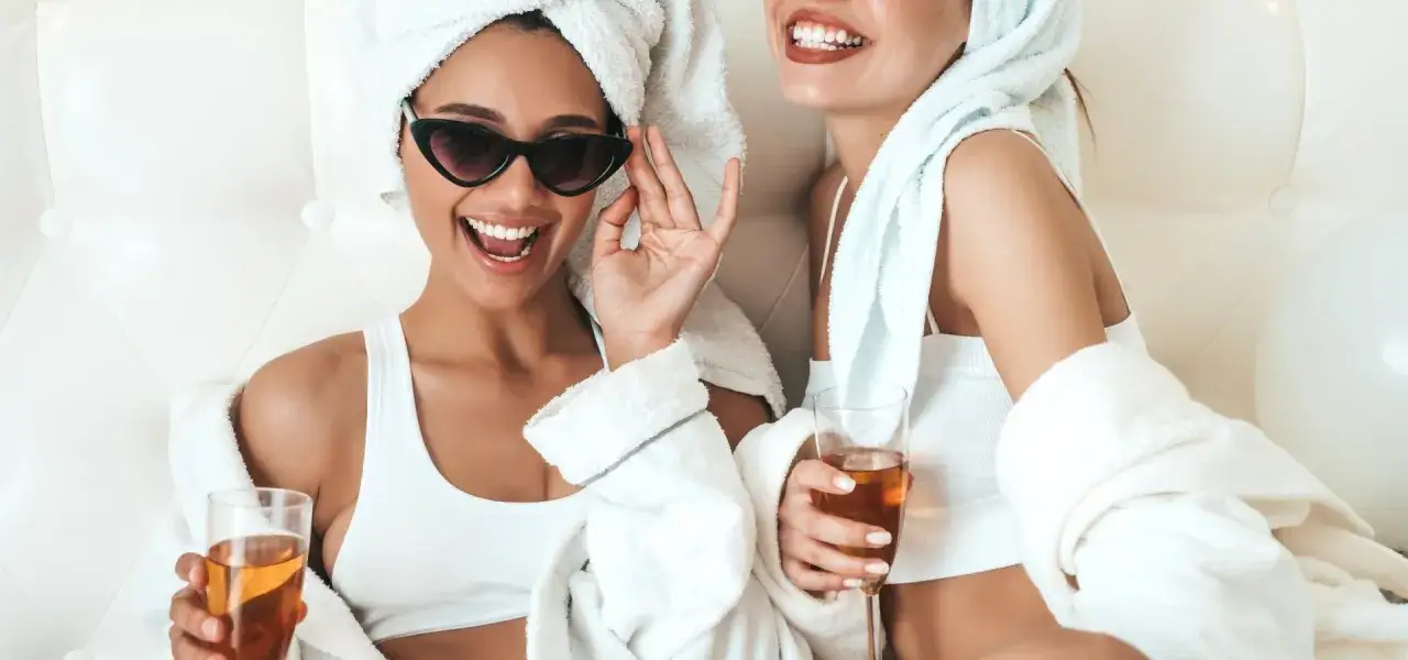Dwie kobiety w szlafrokach i ręcznikach na głowach świętują Dzień Kobiet. Jedna w okularach przeciwsłonecznych, obie z drinkami.