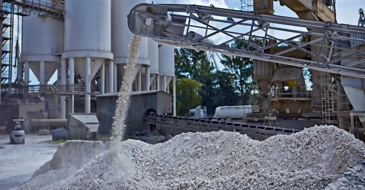 Materiały sypkie, półprodukt do produkcji cementu portlandzkiego, spadają z przenośnika taśmowego na hałdę. W tle silosy i urządzenia przemysłowe.