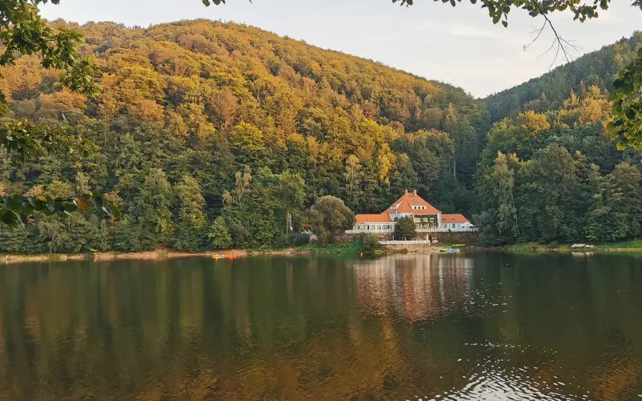 Jezioro otoczone jesiennymi lasami w g&oacute;rach Bystrzyckich. Na brzegu biały budynek z czerwoną dachem, odbijający się w spokojnej wodzie.