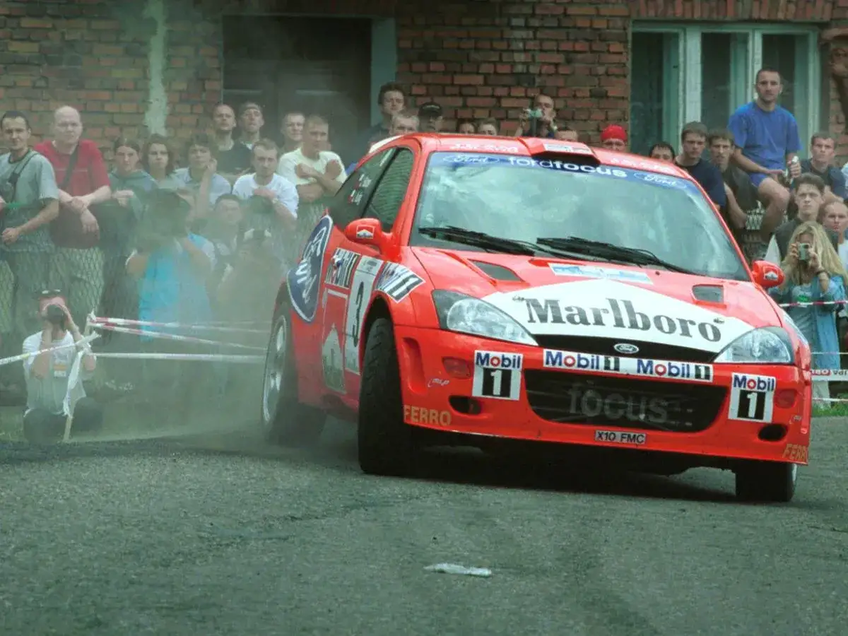 Rajdowy Ford Focus z logiem Marlboro, kierowany przez Janusza Kuliga, w akcji. Widzowie obserwują zmagania.