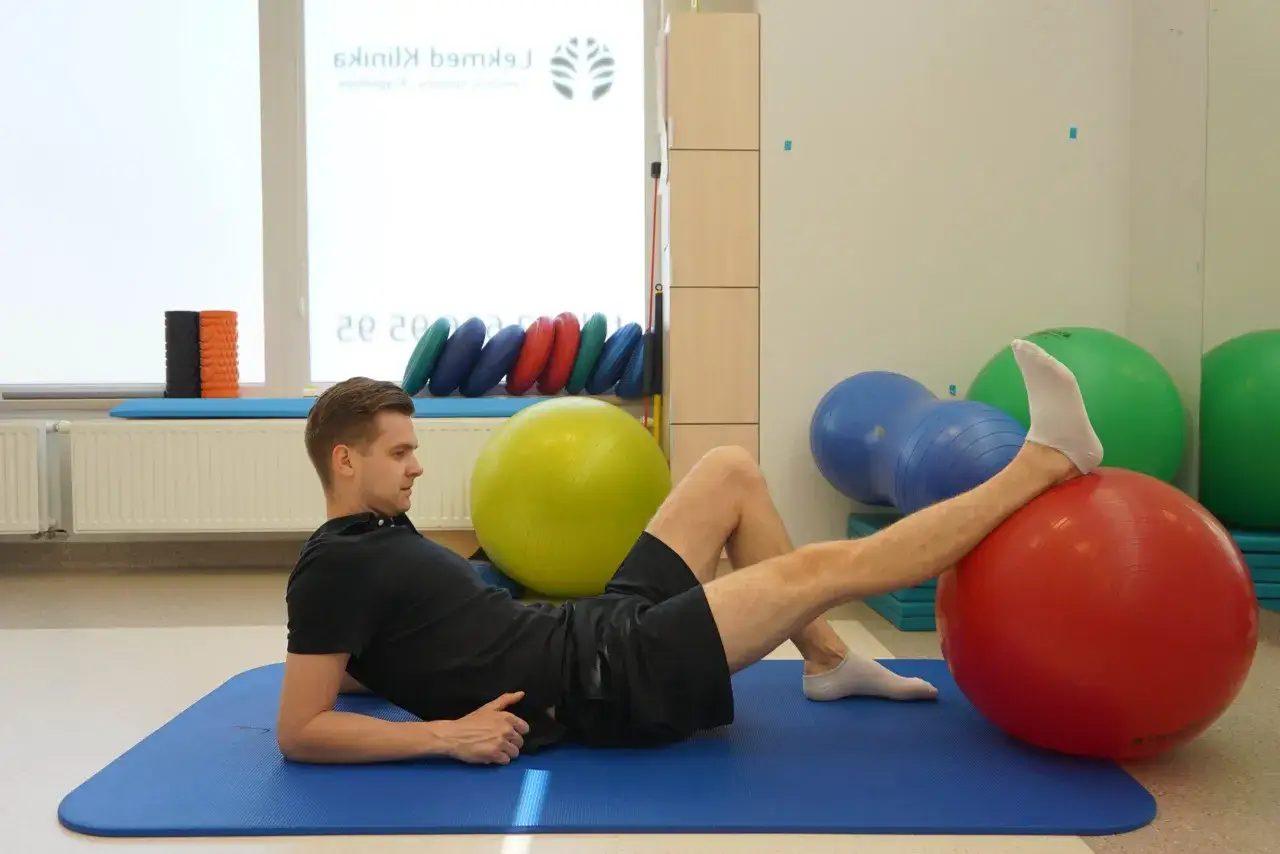 Rehabilitacja po kwasie hialuronowym w kolano: mężczyzna ćwiczy na macie, opierając stopę na czerwonej piłce.