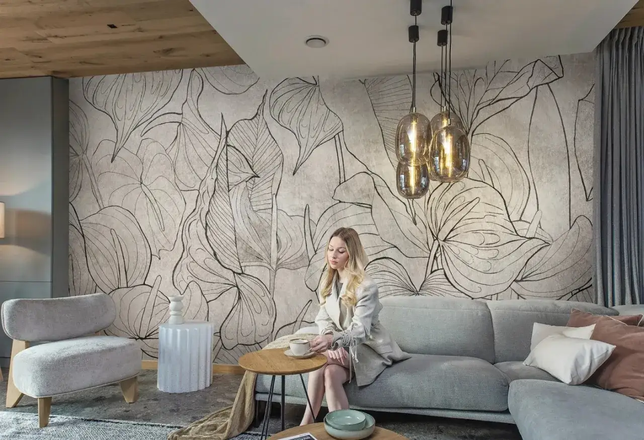 Elegancka kobieta pije kawę na kanapie, podziwiając stylową, tanią tapetę z motywem liści.
