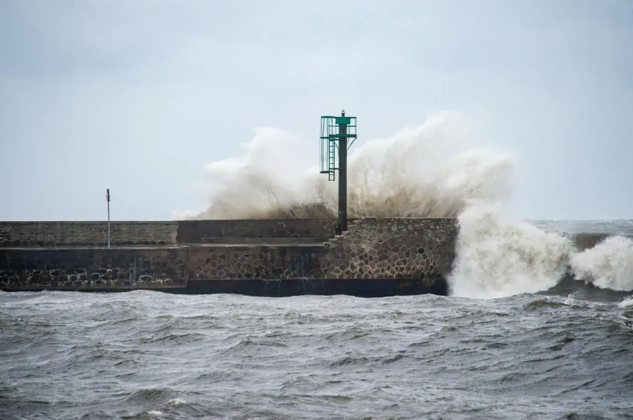 Potężne fale rozbijają się o falochron, kiedy sztormy na Bałtyku szaleją. Latarnia morska stoi niewzruszona.