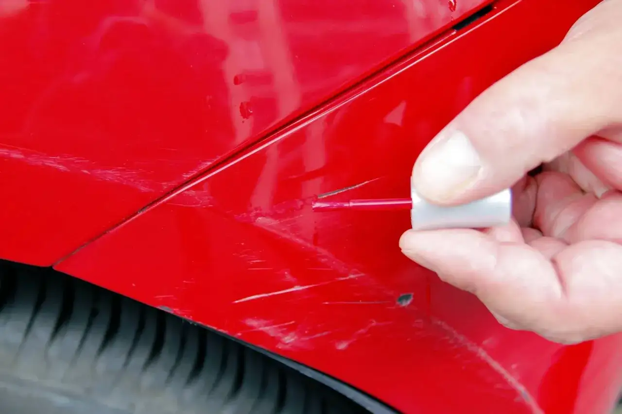 Ręka aplikuje lakier z pędzelka na głębokie rysy czerwonego lakieru samochodu, pokazując, jak usunąć głębokie rysy na lakierze.