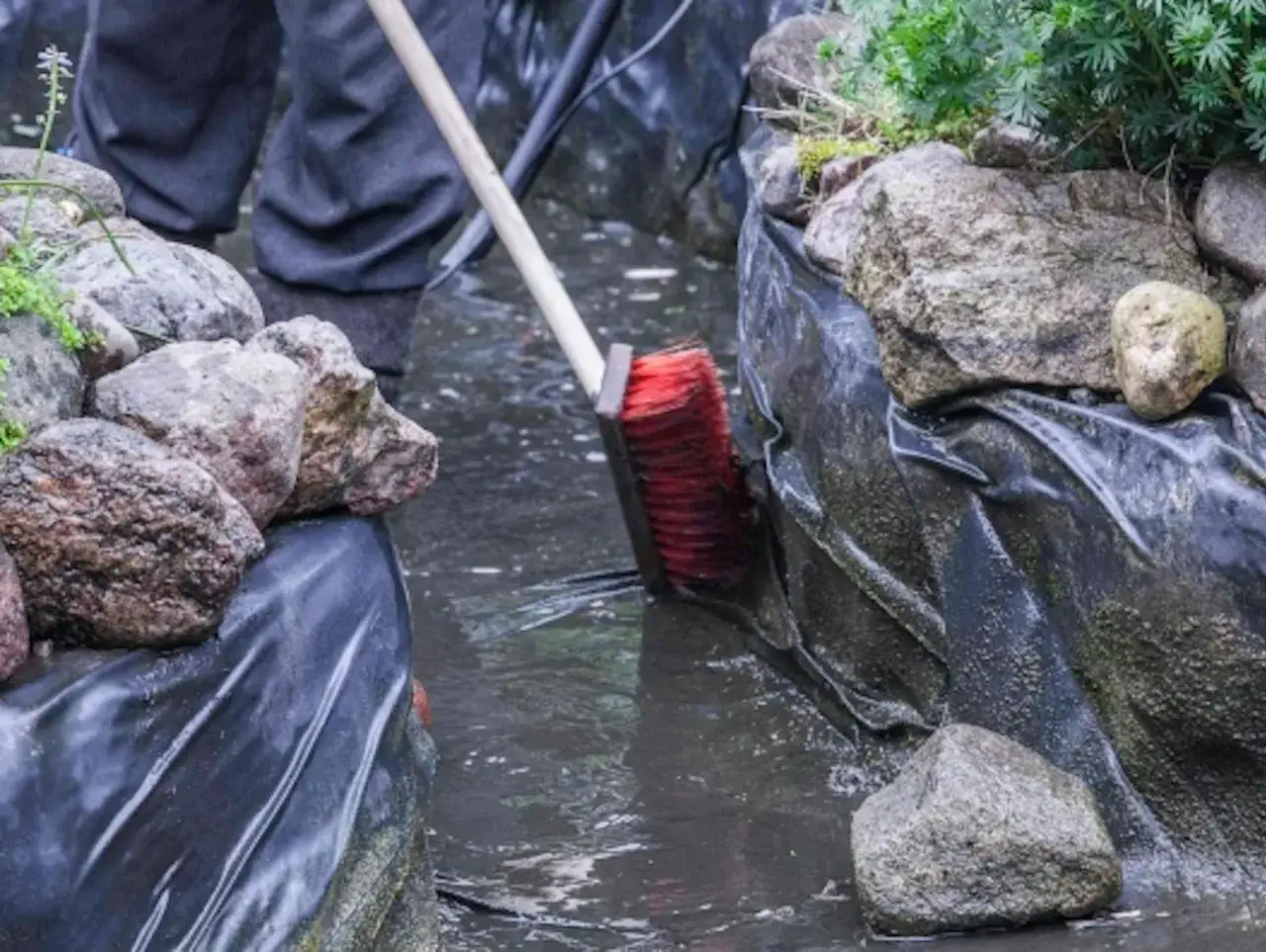 Osoba w gumowcach czyści oczko wodne czerwoną szczotką, usuwając muł i liście.