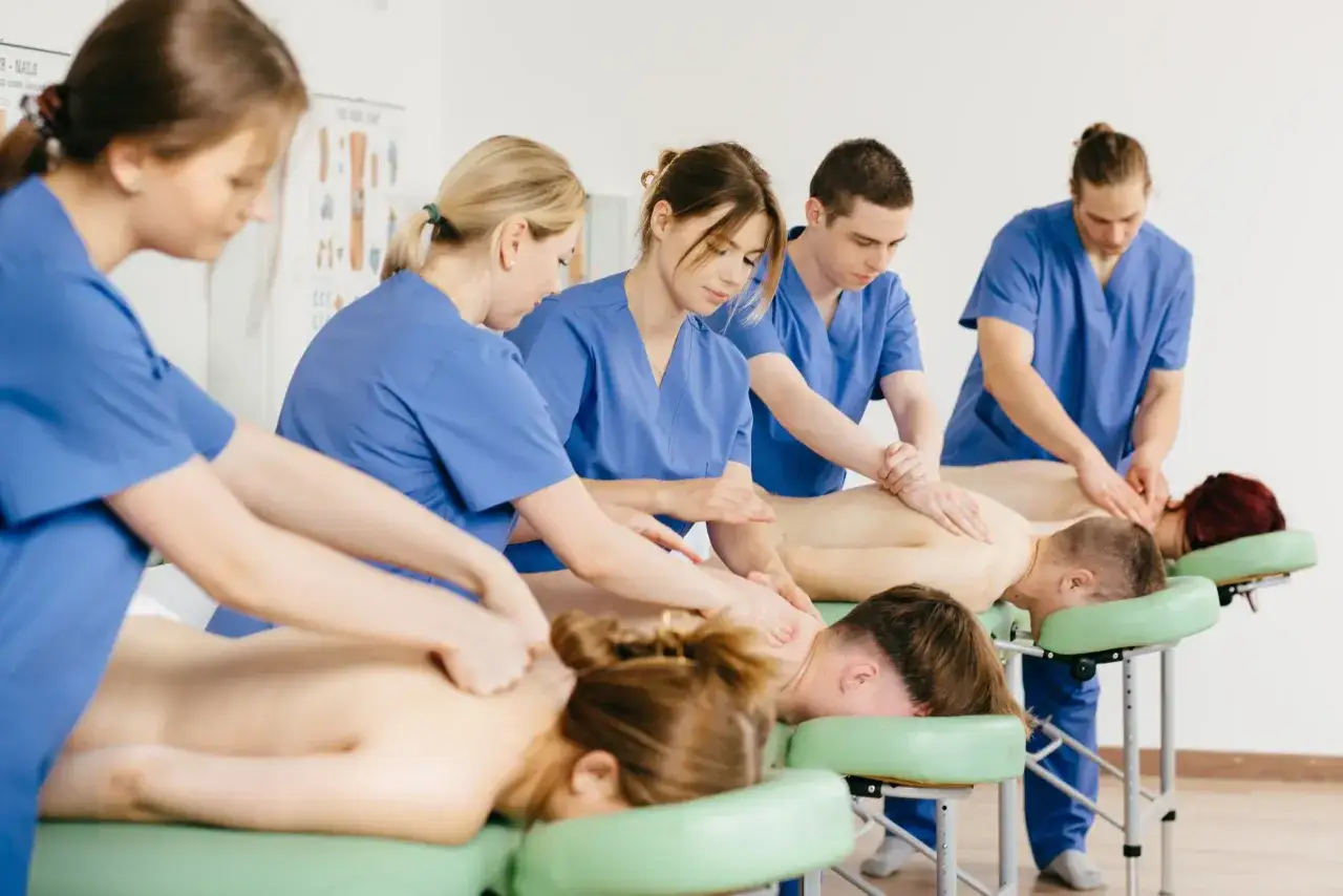 Uczniowie podczas egzaminu zawodowego technik masażysta ćwiczą masaż na leżących na kozetkach osobach.