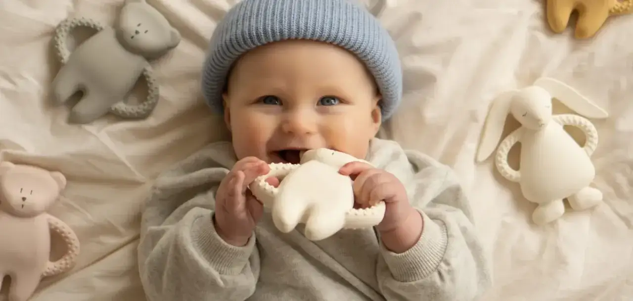 Uśmiechnięty niemowlak w niebieskiej czapce gryzie białą zabawkę-króliczka. Wokół leżą inne gryzaki: miś i szary miś.