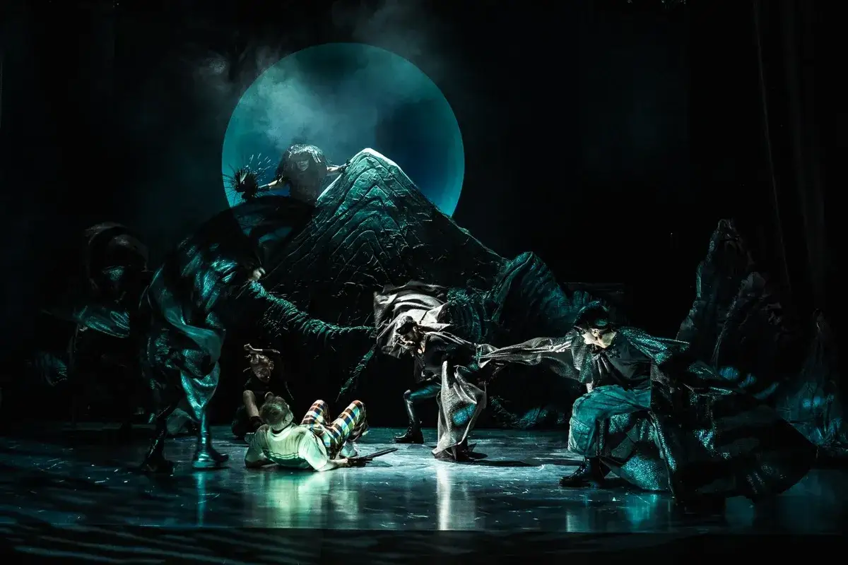 Scena teatralna w Łodzi: mroczna, surrealistyczna wizja z postaciami w fantazyjnych strojach i wielką, niebieską tarczą na tle.
