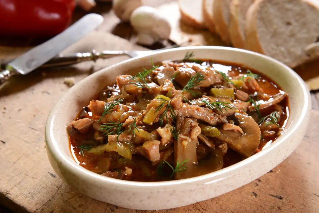 Tradycyjny strogonow z wołowiny i pieczarek, doprawiony koperkiem, podany w ceramicznej misie. Obok kromki chleba i czerwona papryka.