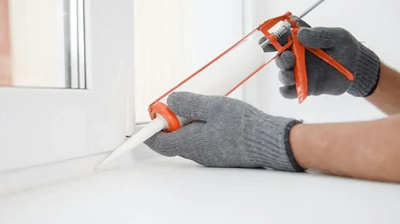 Ręce w rękawiczkach nakładają silikon na ramę okna. Czy silikon można nakładać warstwami? Tak, można, tworząc szczelne połączenie.