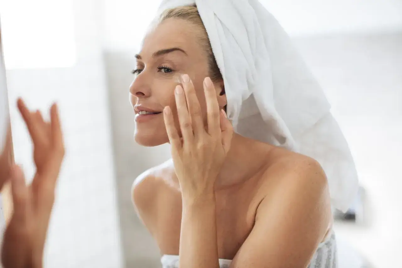 Kobieta z ręcznikiem na głowie aplikuje krem pod oczy. Nawet latem retinol może być Twoim sprzymierzeńcem w walce o gładką sk&oacute;rę.