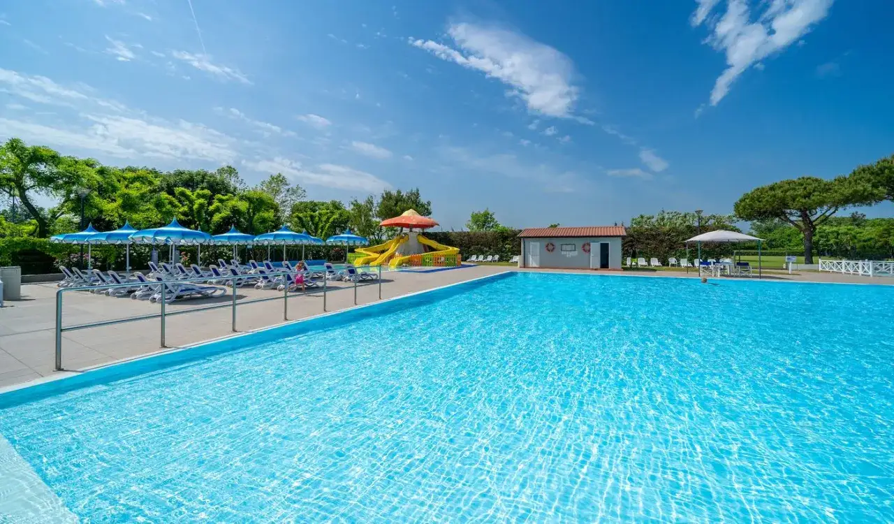 Słoneczny dzień na basenie w Camping Rubicone. Leżaki pod niebieskimi parasolami, zjeżdżalnia dla dzieci i czysta, błękitna woda zapraszają do relaksu.