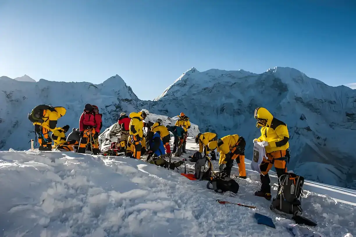 Grupa wspinaczy w ż&oacute;łtych kurtkach przygotowuje się na szczycie island peak. W tle majestatyczne, ośnieżone g&oacute;ry.