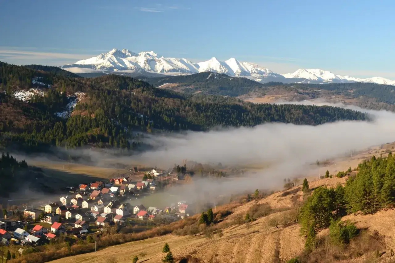 Widok na wieś otoczoną mgłą, zalesione wzg&oacute;rza i ośnieżone szczyty w tle. To malownicze **lesnicke sedlo** o poranku.