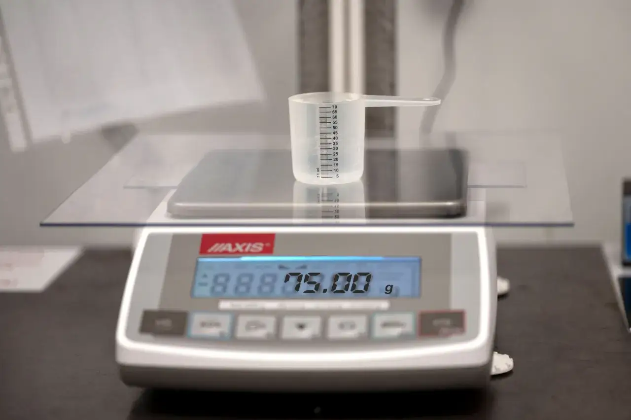 Ważenie proszku na wadze cyfrowej. Miarka z podziałką wskazuje, ile ml ma jedna łyżeczka. Waga pokazuje 75.00 g.