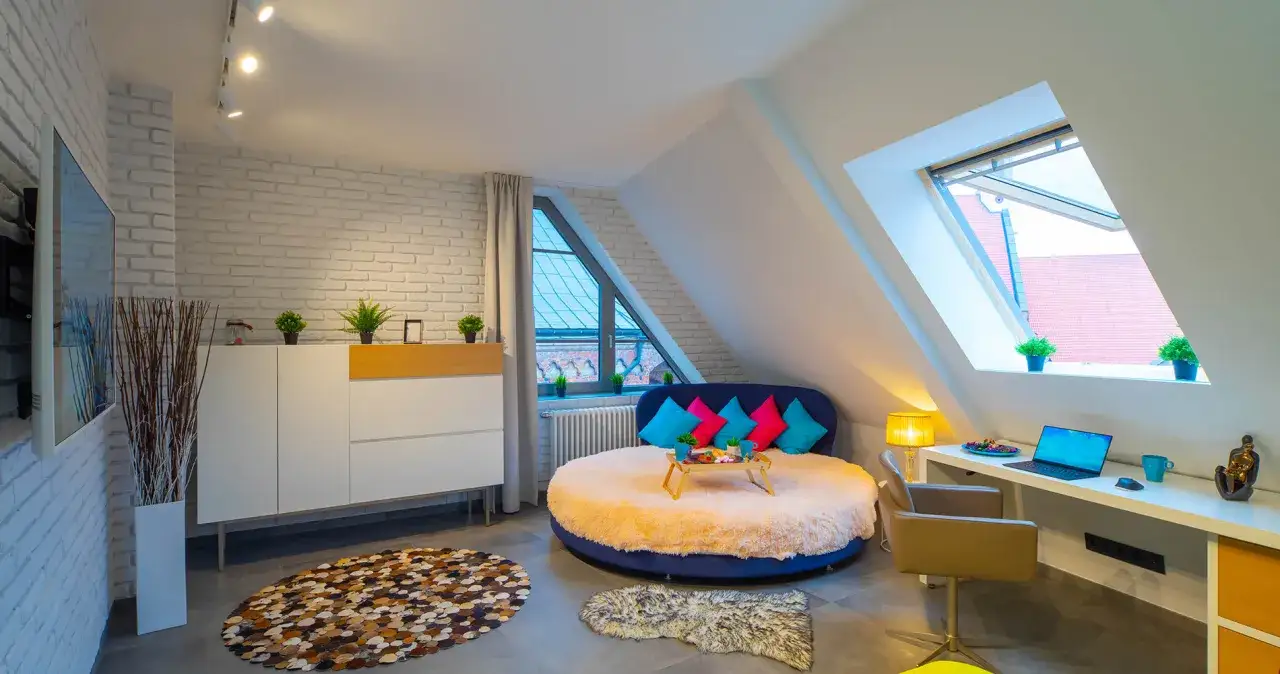 Oświetlenie poddasza: okna dachowe, reflektory i lampka tworzą przytulną atmosferę. Okrągłe łóżko z kolorowymi poduszkami.