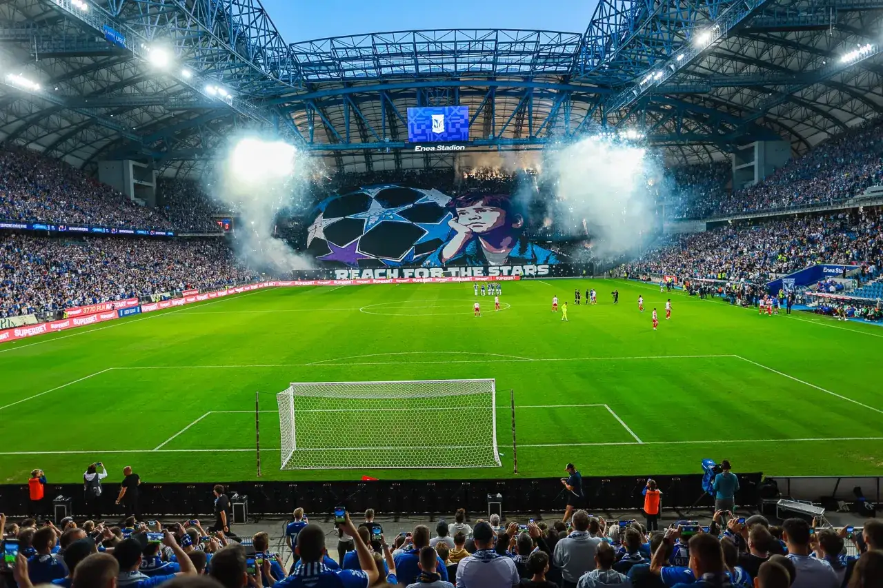 Stadion wypełniony kibicami, dym i pirotechnika. Na telebimie wielka grafika z piłką i hasłem