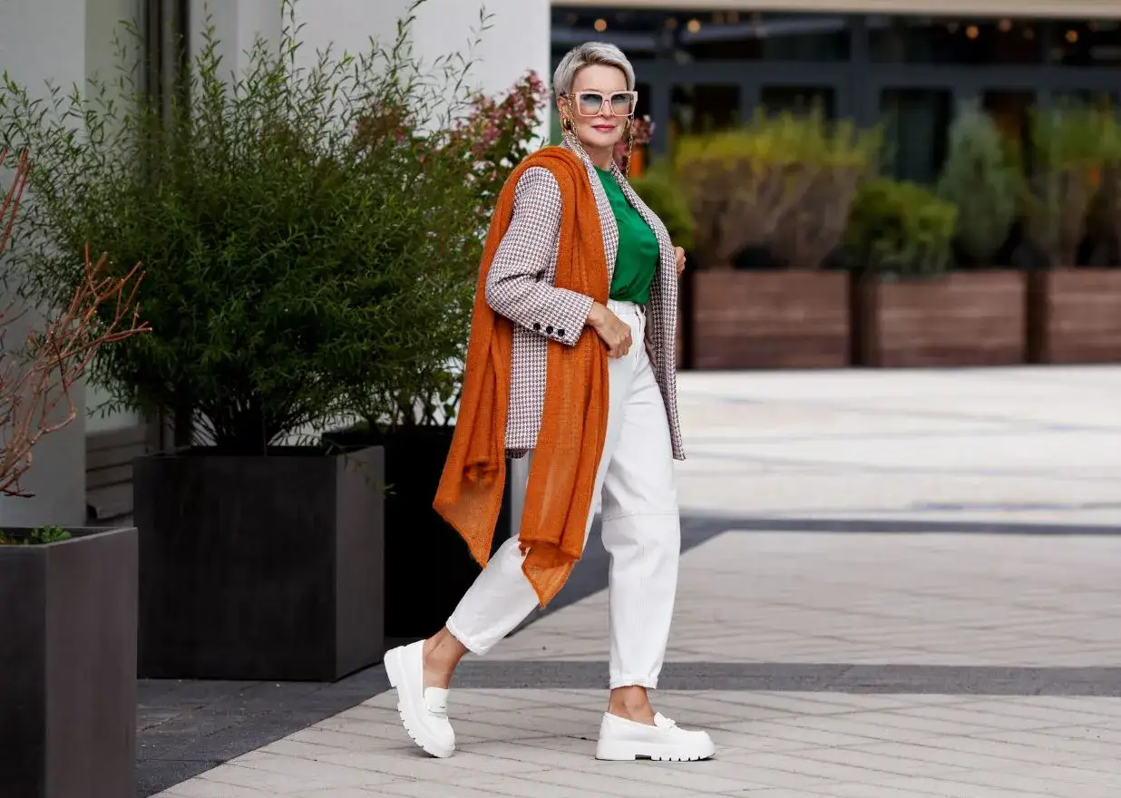 Stylowa kobieta po 50-tce w eleganckim płaszczu, zielonej bluzce i białych spodniach. Pomarańczowy szal dodaje jej szyku.