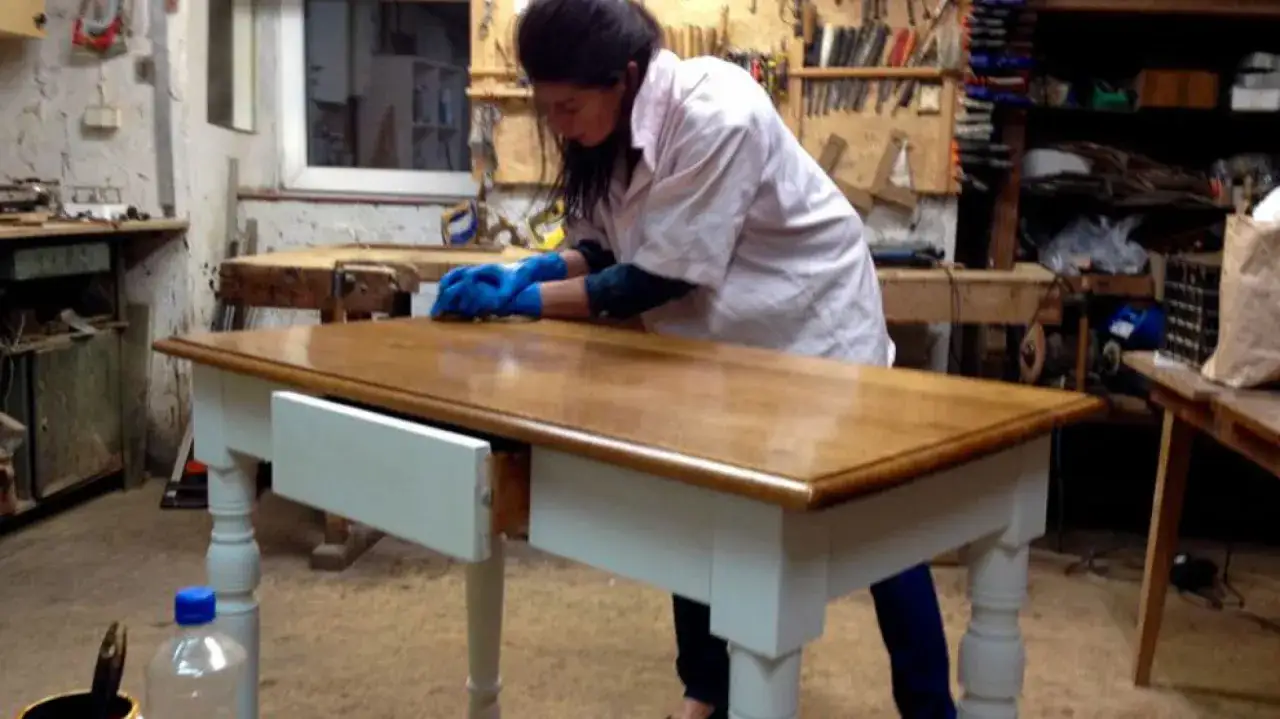 Kobieta w niebieskich rękawiczkach szlifuje blat stołu, kontynuując renowację antyków w warsztacie.