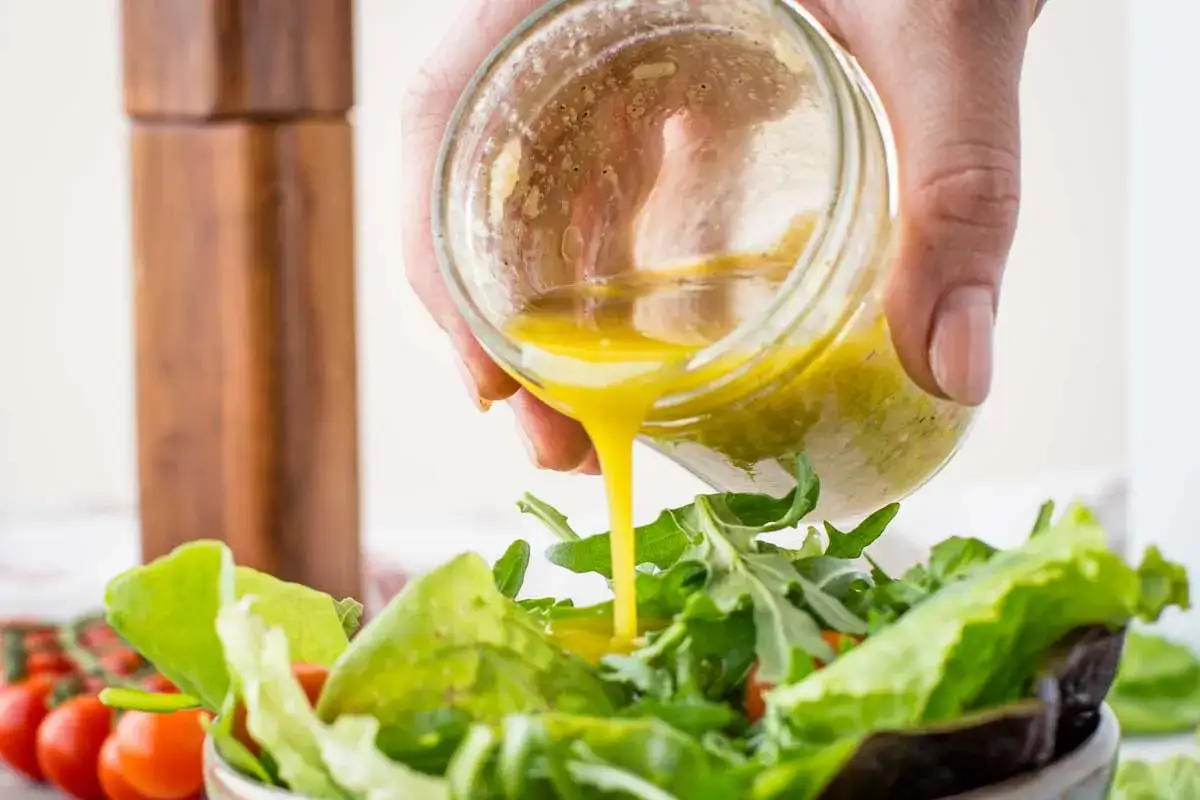 Sos winegret przepis: ręka wylewa złocisty dressing z słoiczka na świeżą sałatkę.