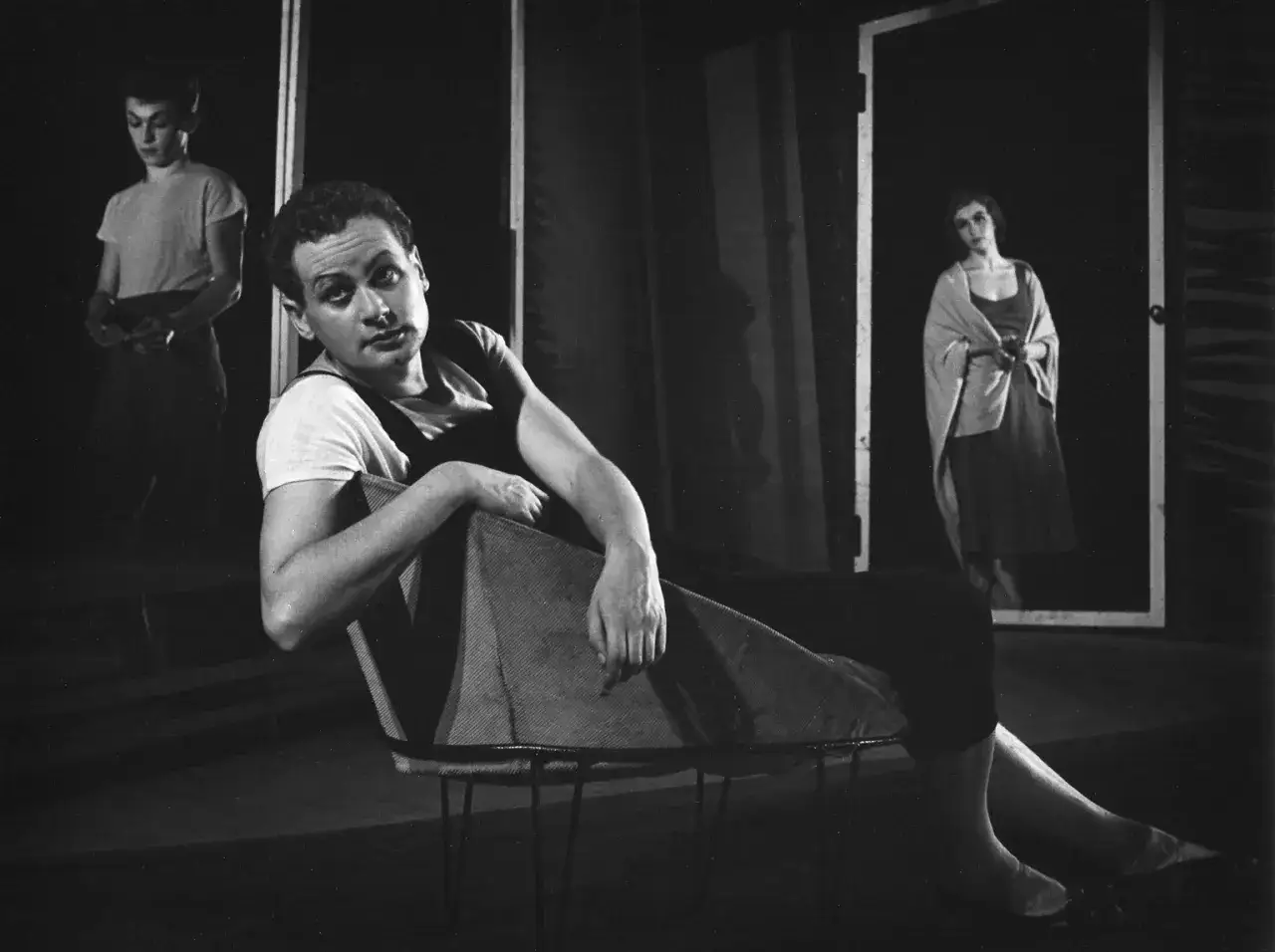 Kto to jest mim? Aktor w białej koszulce i czarnym kombinezonie siedzi na krześle. W tle kobieta w drzwiach i druga postać.