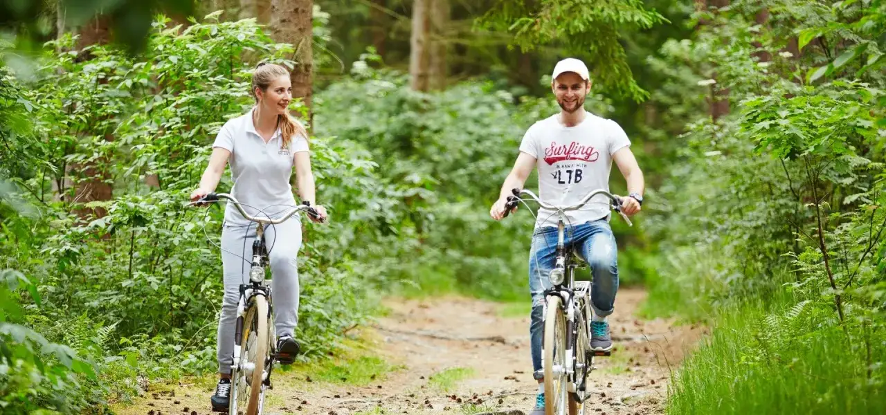 Para na rowerach leśną ścieżką. Ona w białej bluzce, on w koszulce z napisem 