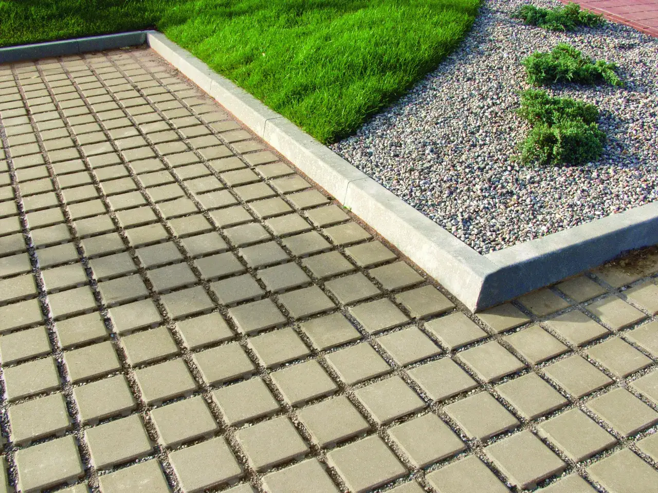 Kostka brukowa przed domem inspiracje: jasny, kwadratowy bruk z żwirkiem między kostkami, obok trawnik i kamyczki.