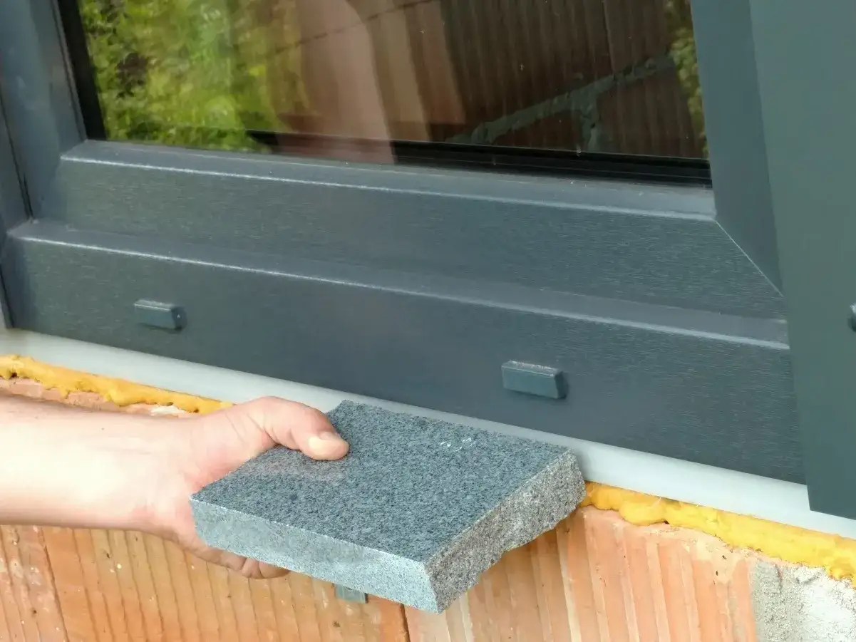 Dłoń trzyma szary kamienny element, który ma być zamontowany jako parapet. Czy parapet jest konieczny? To pytanie o funkcjonalność i estetykę.