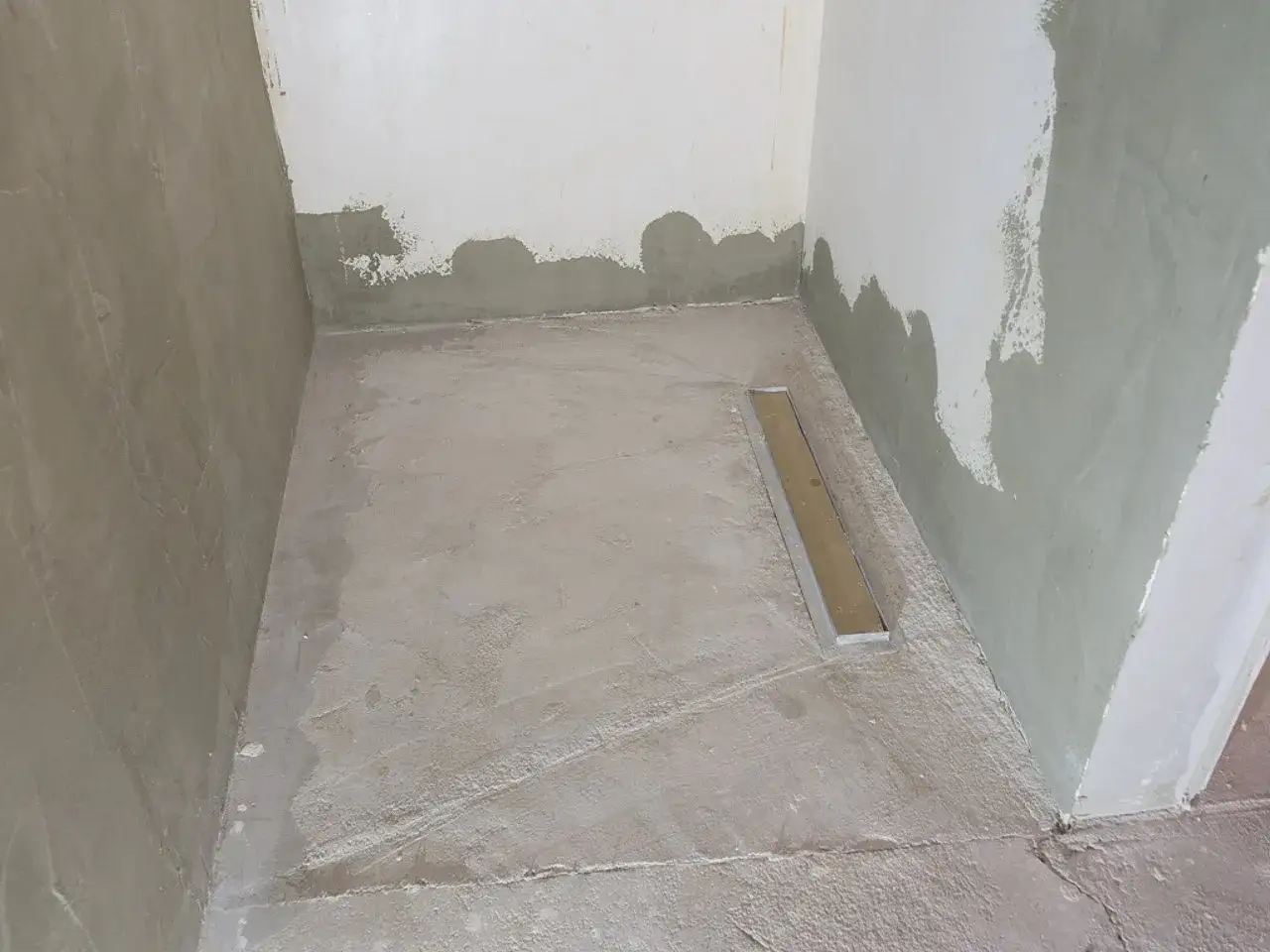 Narożnik łazienki w trakcie remontu. Betonowa podłoga z odpływem liniowym czeka na decyzję: co pierwsze płytki na ścianę czy podłogę?