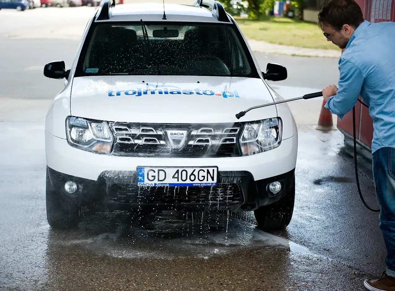Mężczyzna używa myjki ciśnieniowej do obsługi myjni bezdotykowej, spłukując białe Dacia.