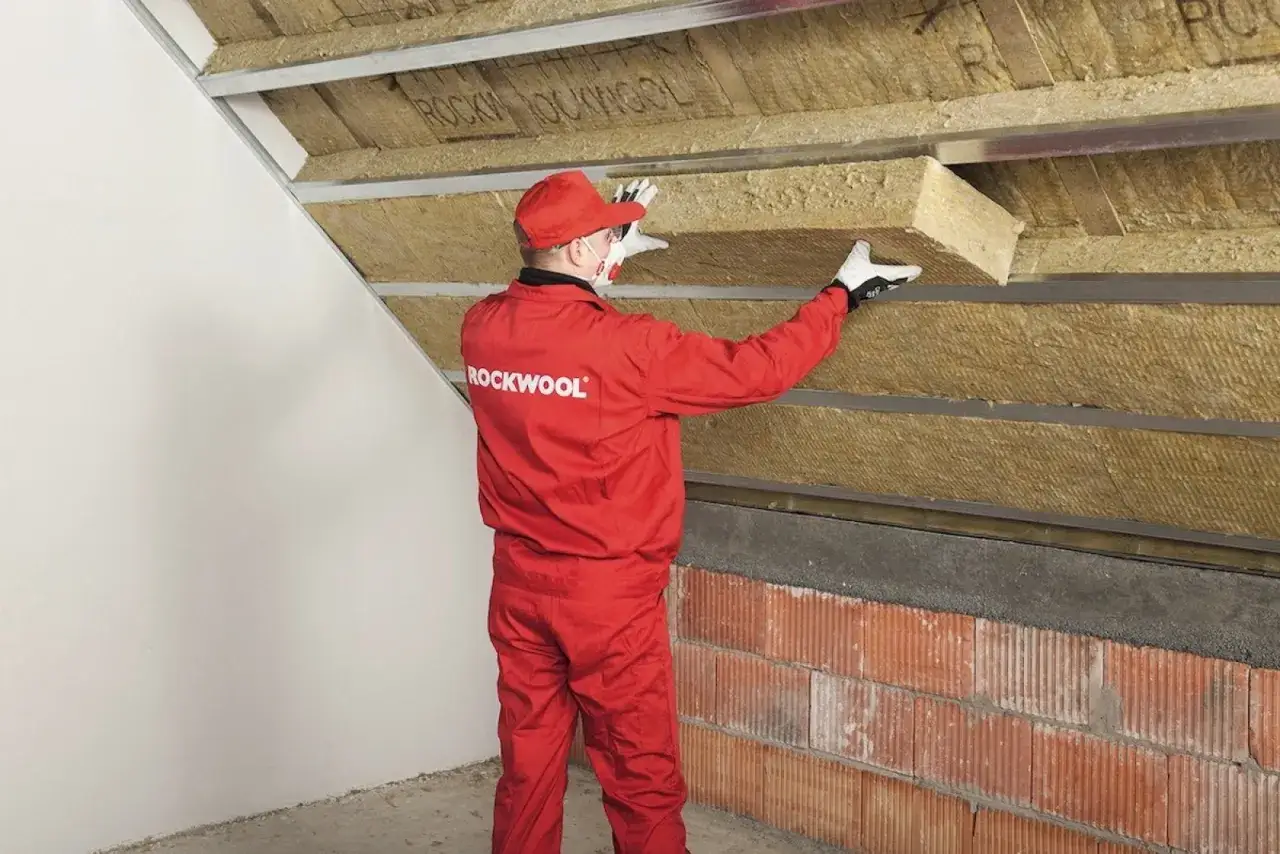 Pracownik w czerwonym kombinezonie montuje wełnę mineralną, wykonując wykończenie ściany szczytowej poddasza.