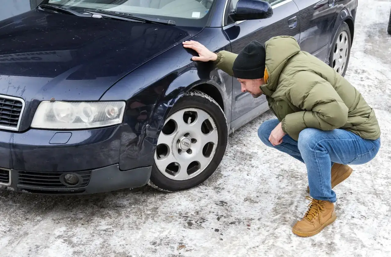 Mężczyzna w zimowej kurtce klęczy przy ciemnym samochodzie, oglądając nieświadome zarysowanie na błotniku.