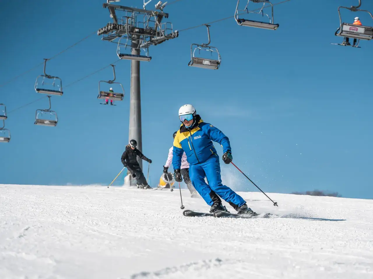 Narciarz w niebiesko-żółtej kurtce szusuje po stoku na ski station Suche. W tle widać wyciąg krzesełkowy i innych narciarzy.