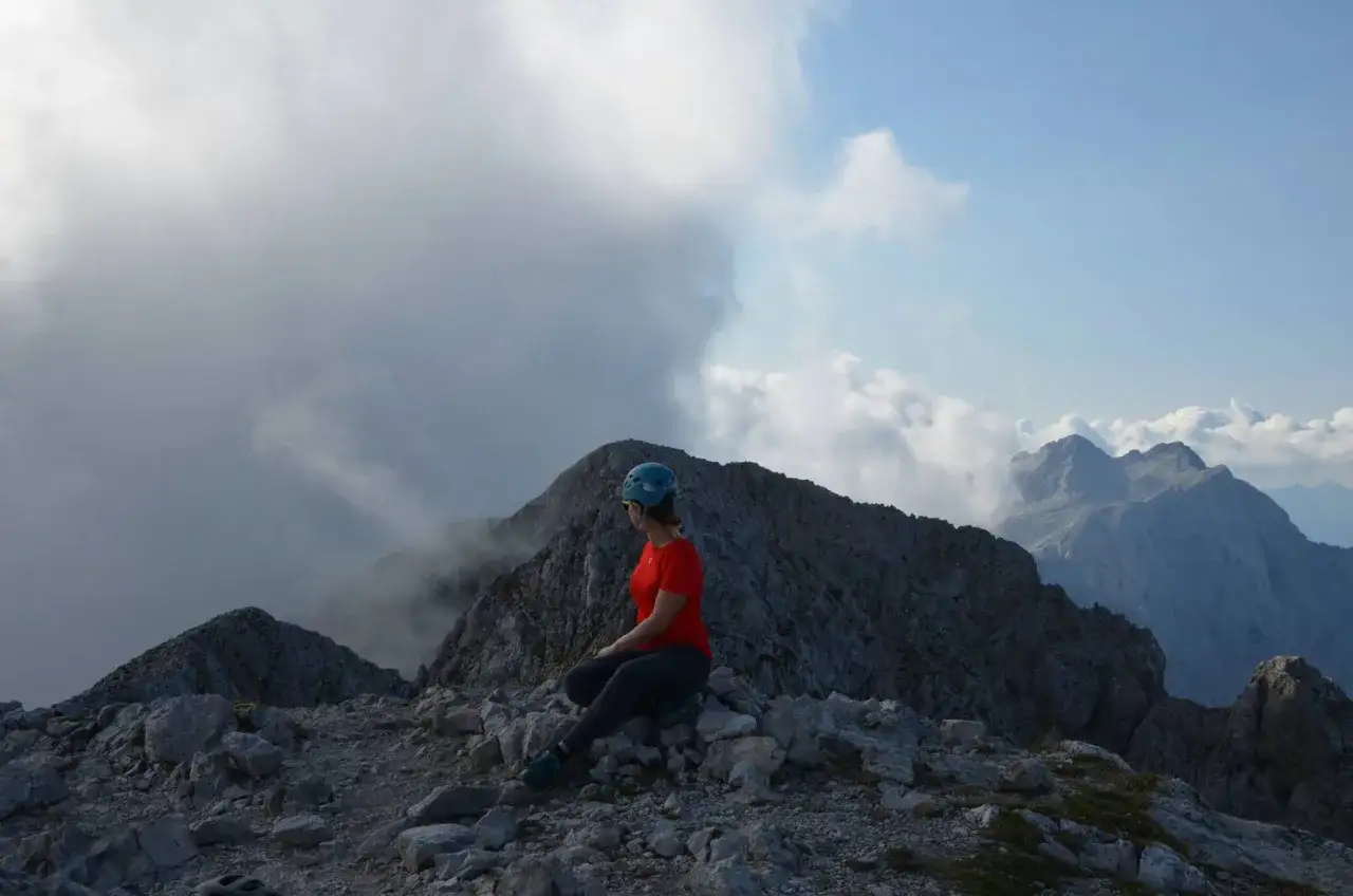 Kobieta w kasku i czerwonej koszulce siedzi na skalistym szczycie, podziwiając majestatyczne **Alpy Kamnickie** i chmury.