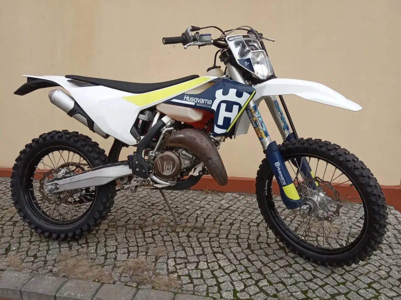 Husqvarna 125 Enduro gotowy do akcji na brukowanej drodze. Biało-niebieski motocykl z żółtymi akcentami.