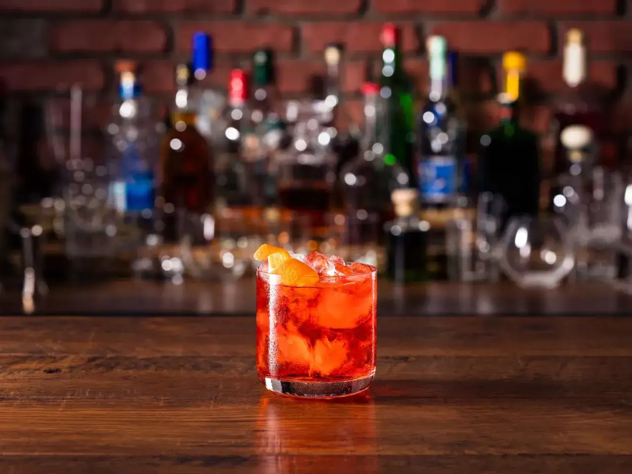 Klasyczny negroni, idealny na wieczór. Czerwony drink z pomarańczą i lodem na drewnianym blacie, w tle bar z butelkami.