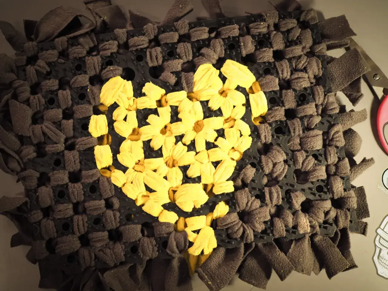 Jak zrobić matę węchową dla psa: czarna podstawa z otworami, w kt&oacute;rą wpleciono szare i ż&oacute;łte paski materiału, tworząc serce.