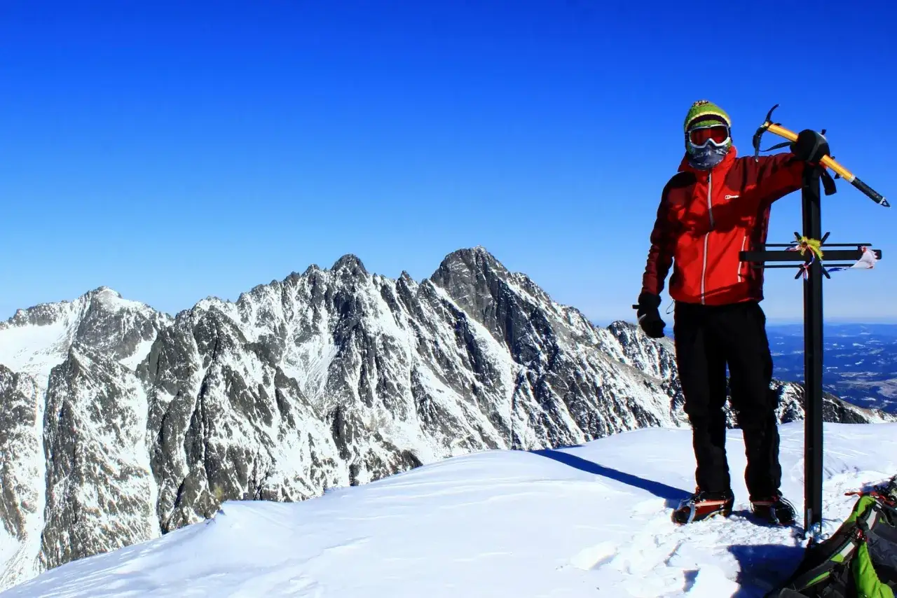 Wspinacz na szczycie w Tatrach w maju, z lodowym toporem opartym o krzyż. W tle ośnieżone szczyty i błękitne niebo.