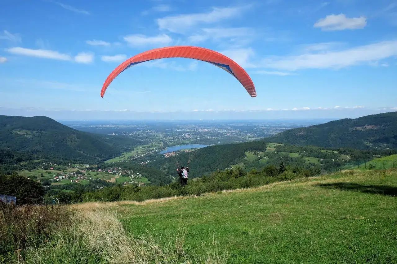 Paralotniarz szybuje nad zieloną doliną, gdzie jest góra Żar. W oddali jezioro i miasteczko.