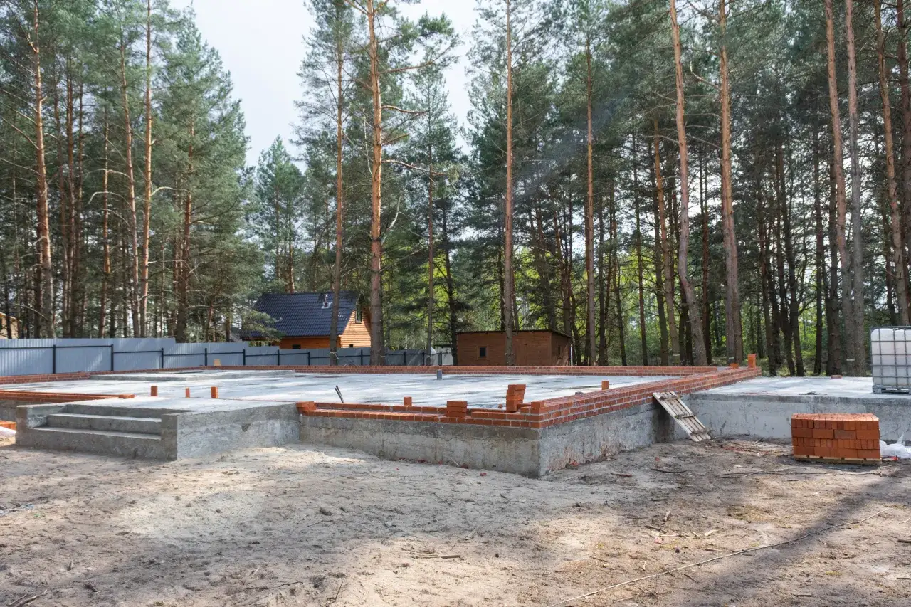 Fundament domu modułowego w lesie. Widać betonową płytę, schody i początek murowania ścian z cegły.