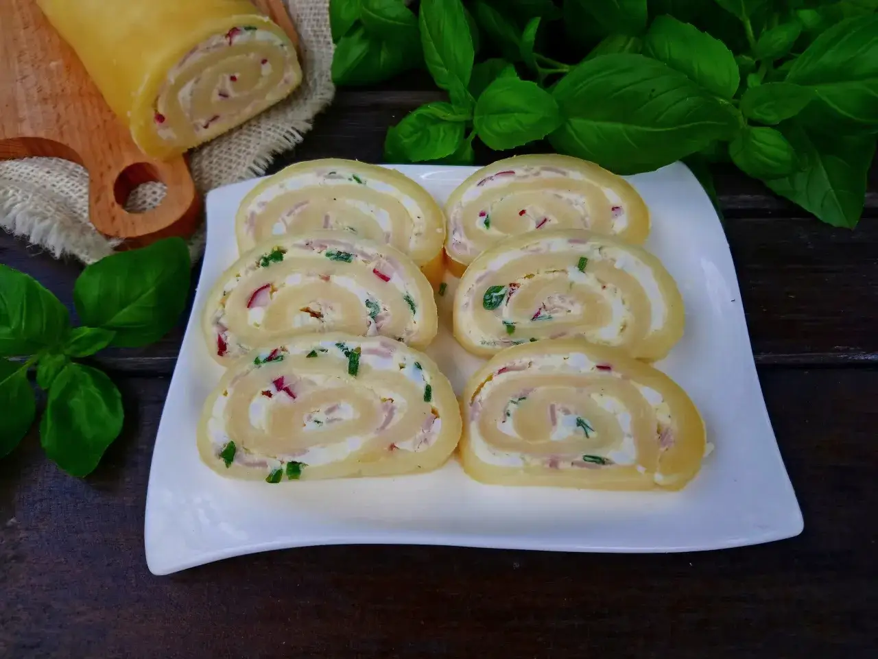 Rolada serowa z wędzonym łososiem, jajkiem i szczypiorkiem, pokrojona na plastry, prezentuje się apetycznie na białym talerzu.