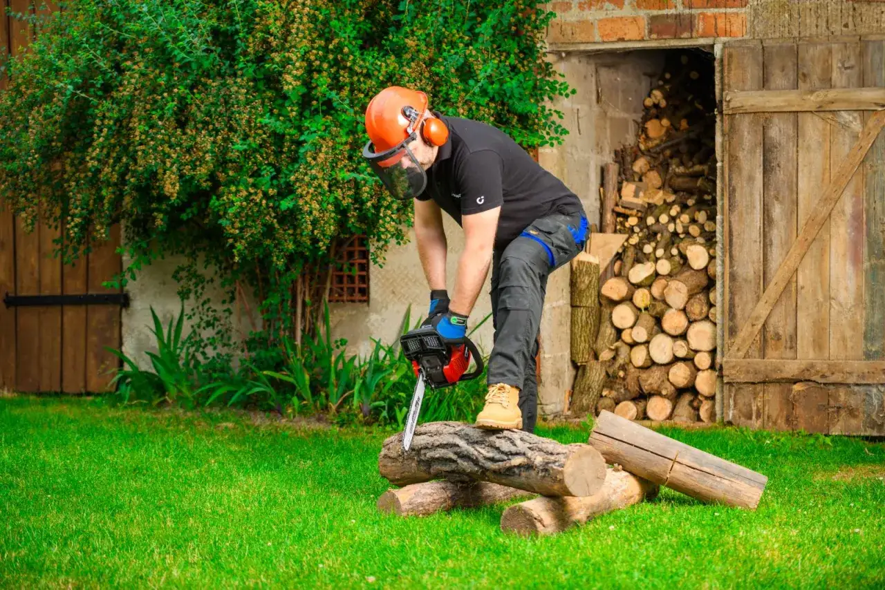 Mężczyzna w kasku i ochronach demonstruje, jak ciąć piłą łańcuchową drewno. W tle sterta drewna i stara stodoła.