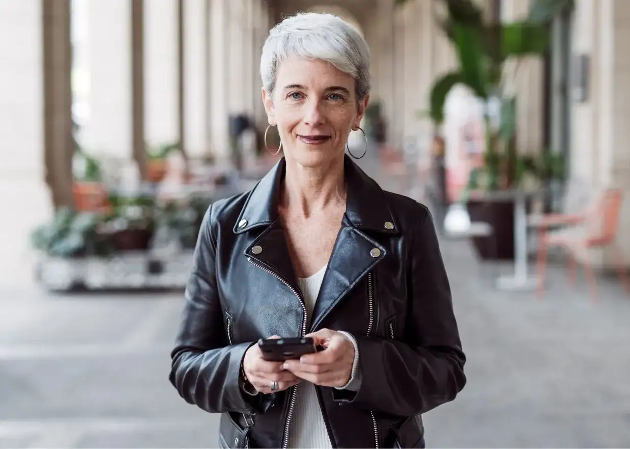 Elegancka kobieta po 50-tce w skórzanej ramonesce, z krótkimi siwymi włosami i okrągłymi kolczykami, trzyma telefon. To inspiracja dla mody dla 50 latki.