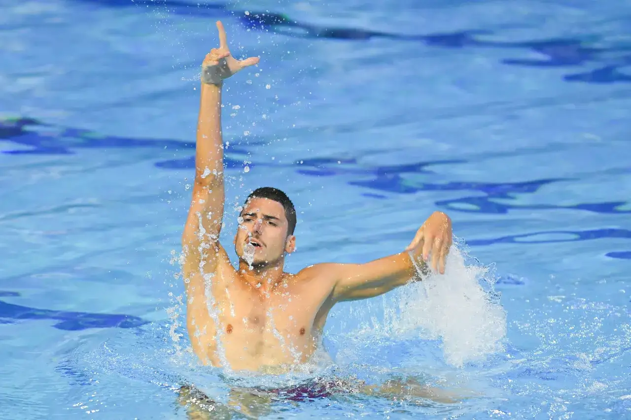 Mężczyzna wykonuje element pływania synchronicznego, unosząc rękę nad wodą, kt&oacute;ra rozpryskuje się wok&oacute;ł.