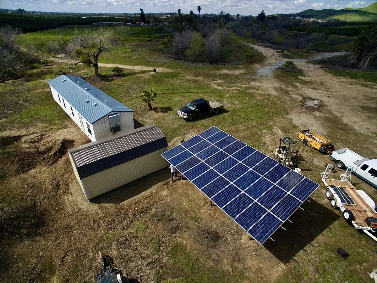 Duży panel słoneczny, tworzący siatkę, stoi obok budynk&oacute;w i pojazd&oacute;w na słonecznym terenie.