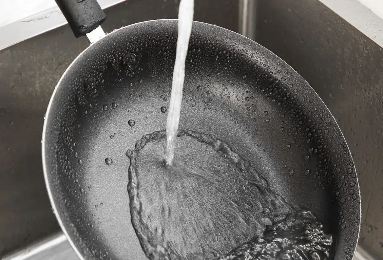 Czyszczenie patelni sodą i octem: strumień wody spływa po wewnętrznej stronie czarnej patelni w zlewie, tworząc pianę.
