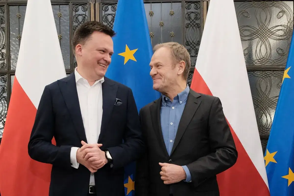 Dwóch liderów partii politycznych, Donald Tusk i Szymon Hołownia, uśmiecha się do siebie na tle flag Polski i Unii Europejskiej.