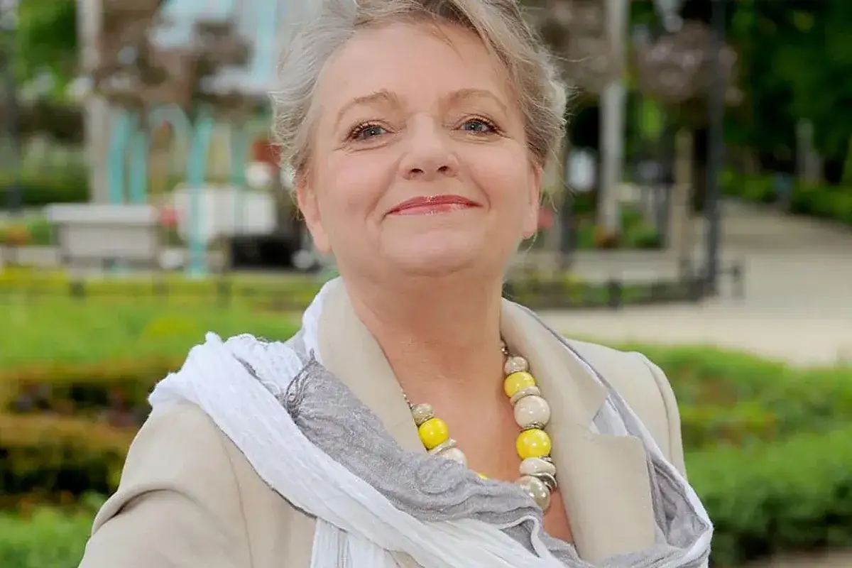 Uśmiechnięta Anna Seniuk, znana z wielu film&oacute;w, seriali i program&oacute;w, w beżowej marynarce i z kolorowym naszyjnikiem.