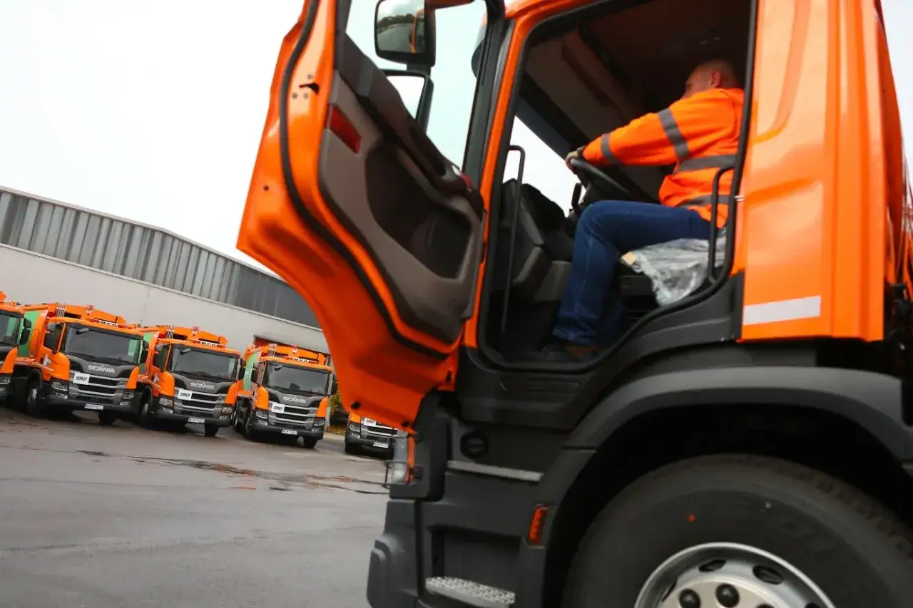 Kierowca śmieciarki w Niemczech w pomarańczowej kamizelce siedzi za kierownicą. W tle rząd pomarańczowych śmieciarek.